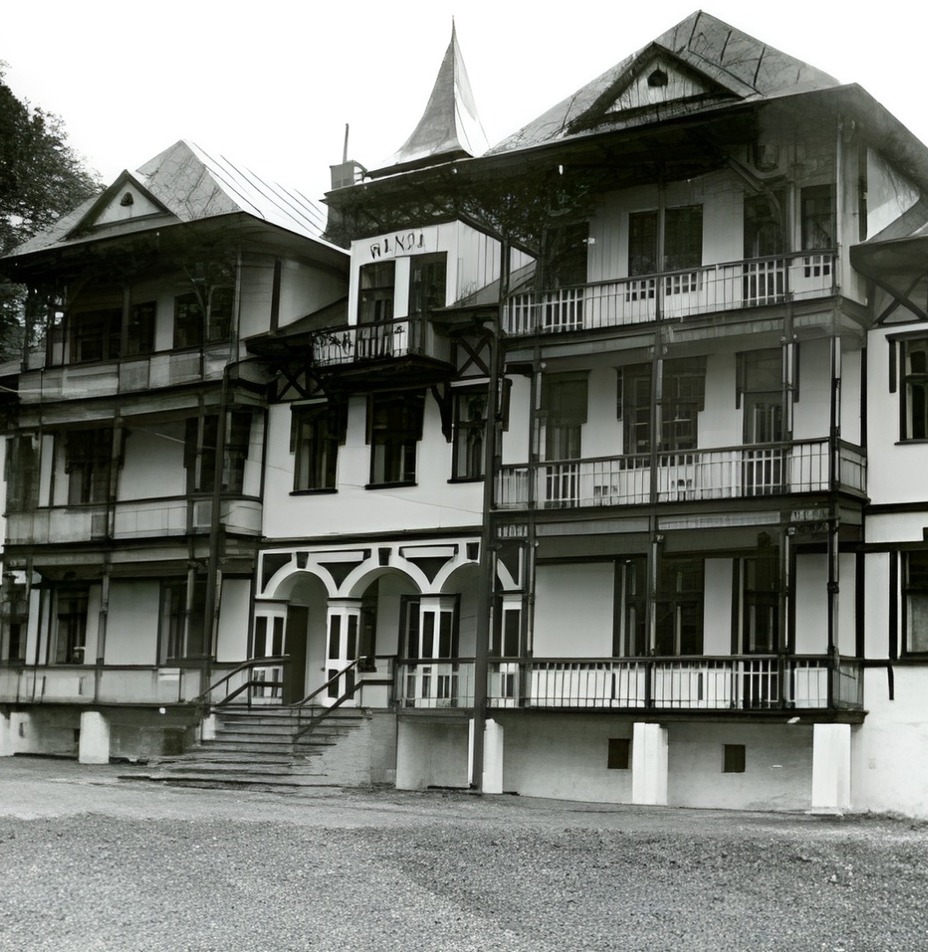 Zabytkowy górski hotel z drewnianymi balkonami i zadaszonymi werandami, wielopiętrowa elewacja na tle zalesionego stoku.