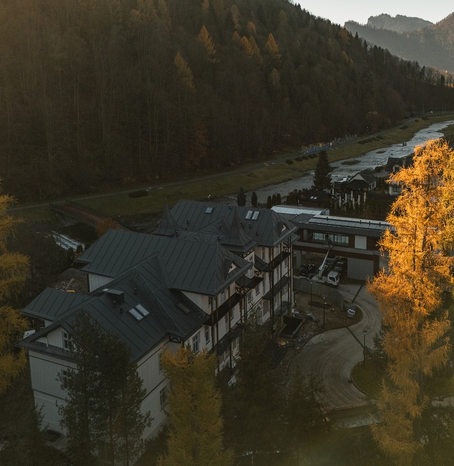 Hotel w stylu alpejskim z ciemnymi dachami i balkonami przy rzece, złote jesienne drzewa w górskiej dolinie o zachodzie słońca