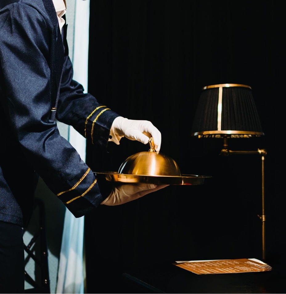 Uniformowany kamerdyner w białych rękawiczkach podaje złotą kopułę na tacy obok mosiężnej lampy