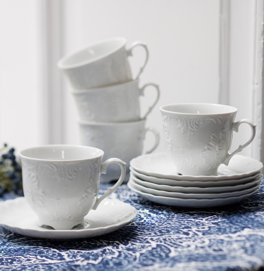 Eleganckie, tłoczone białe filiżanki i spodeczki ułożone na niebieskim wzorzystym obrusie przy jasnym oknie