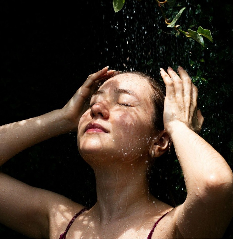 Gość relaksuje się pod zewnętrznym prysznicem deszczowym, dłonie na głowie, otoczony bujną zielenią.