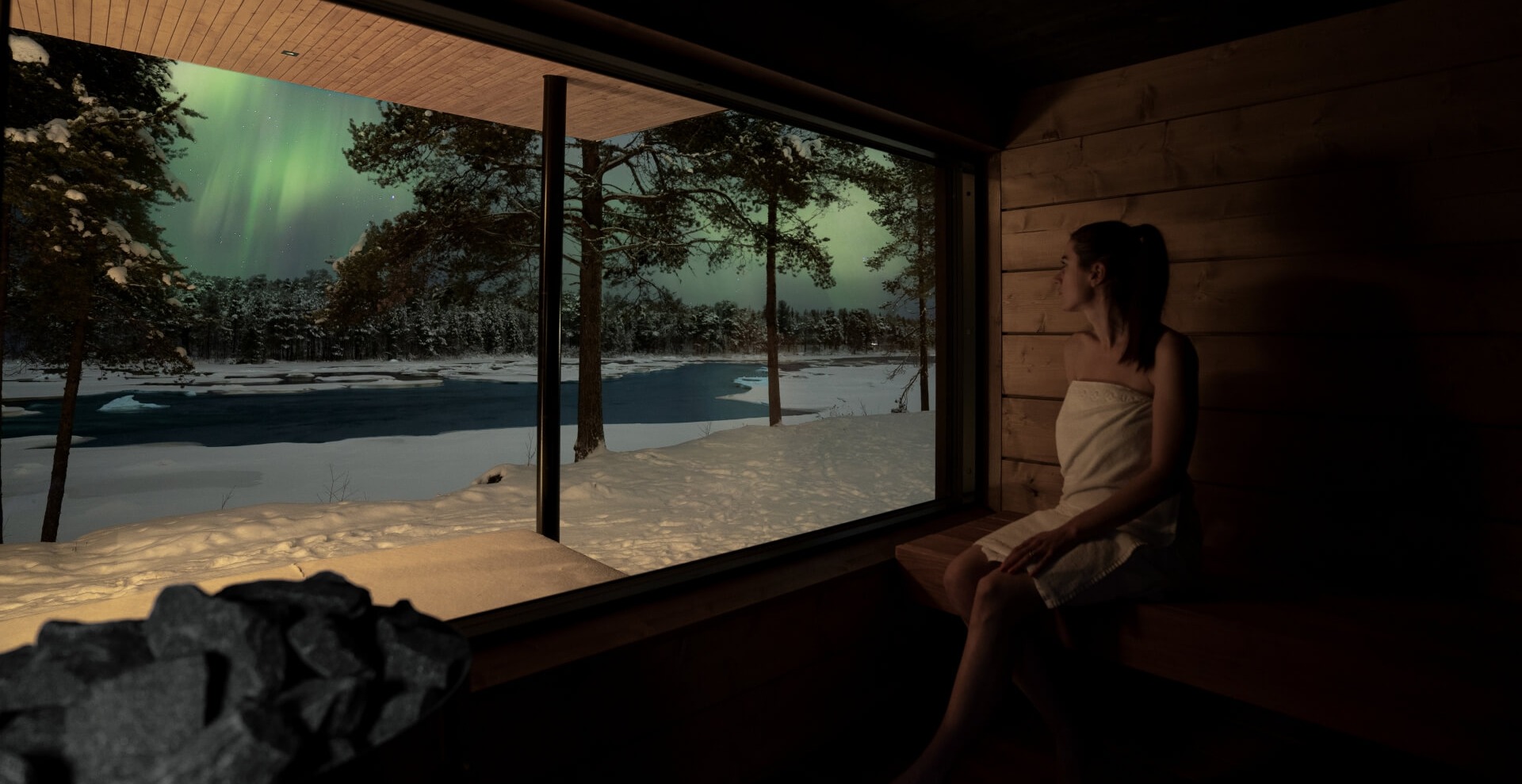Sauna privé en bois avec banquette ; cliente en serviette admire l'aurore boréale au‑dessus d'une rivière enneigée par une grande fenêtre.