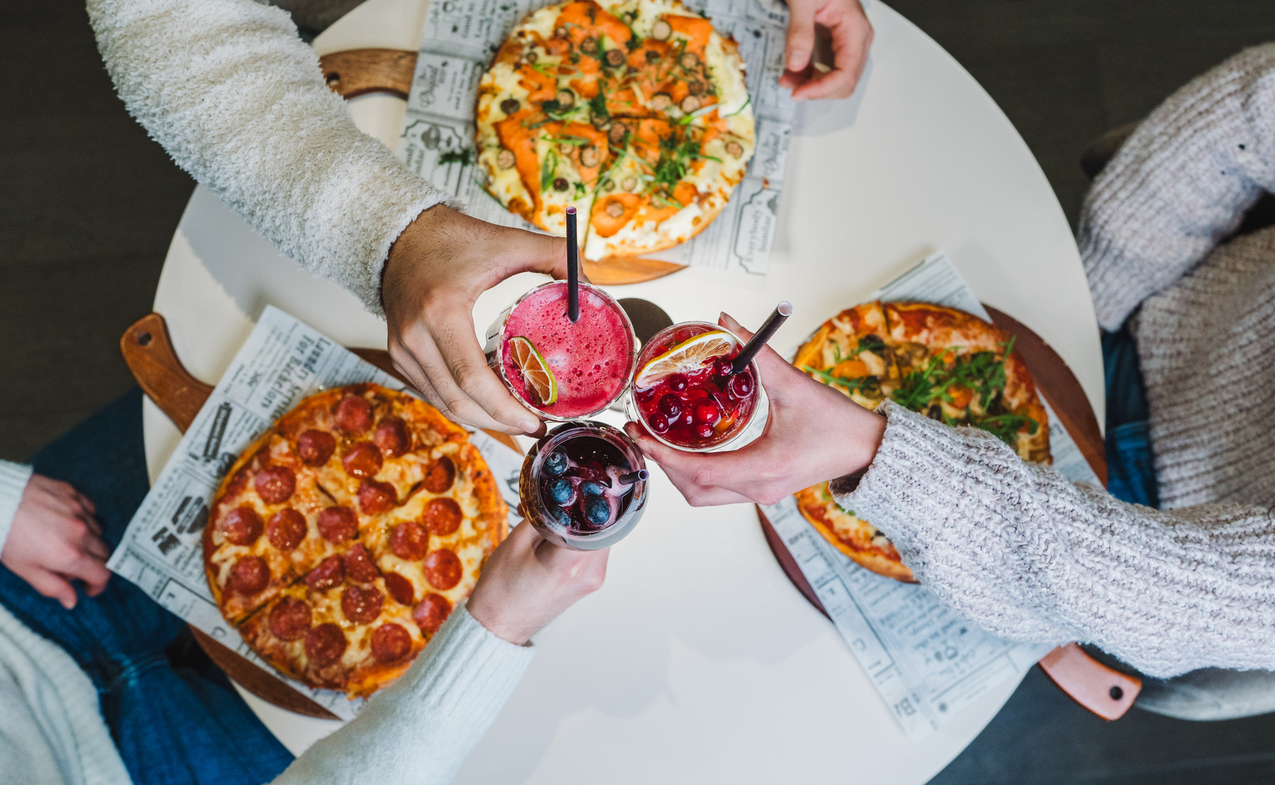 Friends toasting colorful cocktails over a round table with four pizzas on wooden boards, casual shared dining.