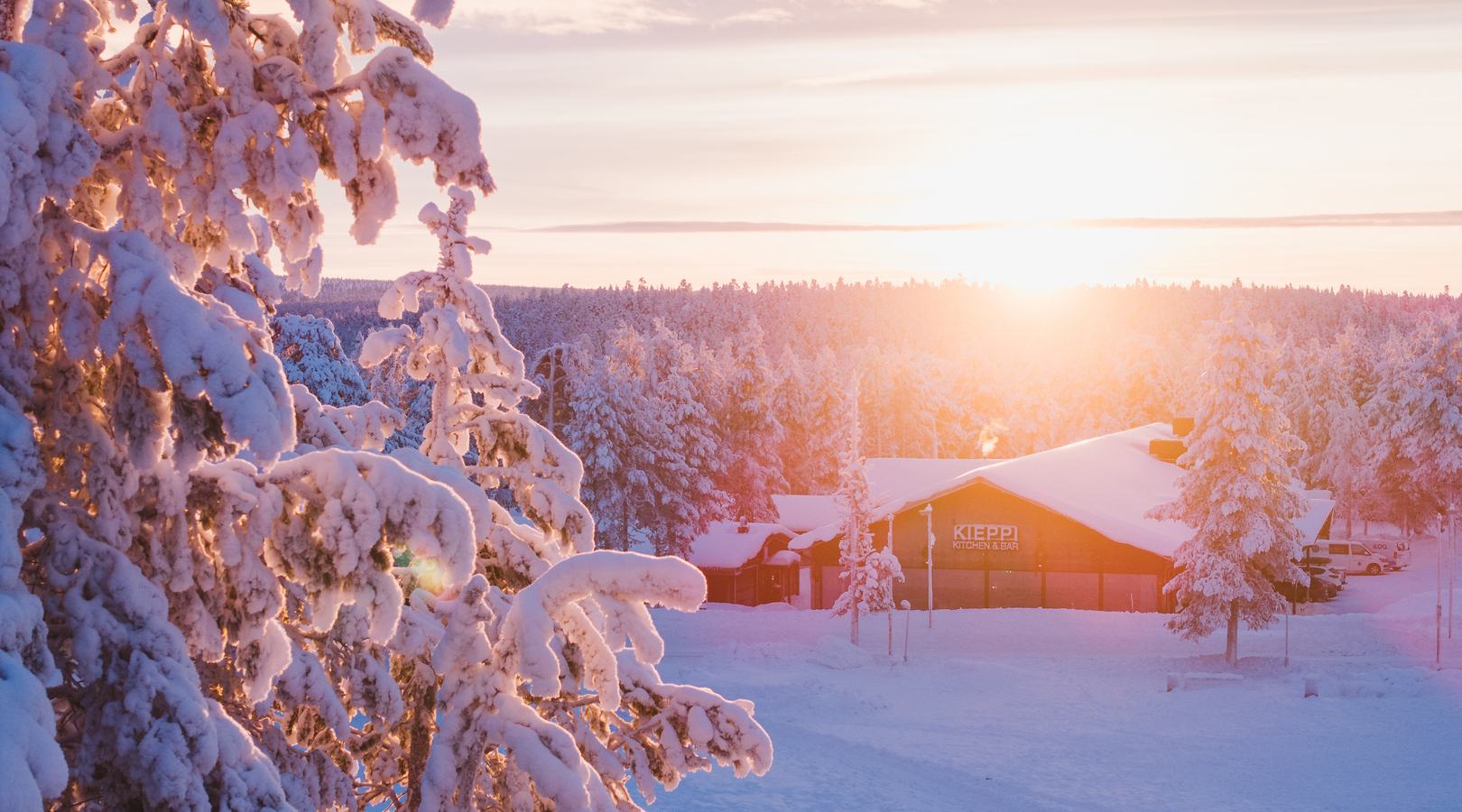 Snow-covered pines and a cozy lodge marked Kieppi Kitchen & Bar, bathed in warm sunrise light across a peaceful winter resort