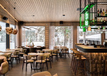 Cozy hotel bar and dining room with wooden ceiling, plaid chairs, round tables and large windows revealing snow-covered forest views.