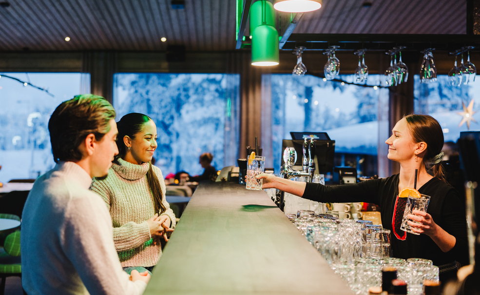 Bar d'hôtel chaleureux où la barmaid sert des cocktails à deux clients au comptoir en bois, lumières douces et vue enneigée