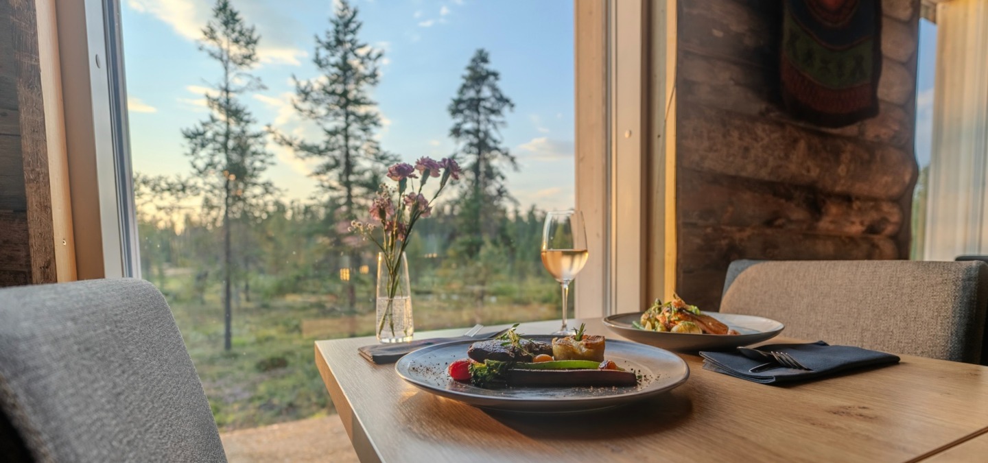 Wood-paneled dining table by a large window with plated gourmet dishes, a glass of white wine, fresh flowers and a peaceful pine-forest view