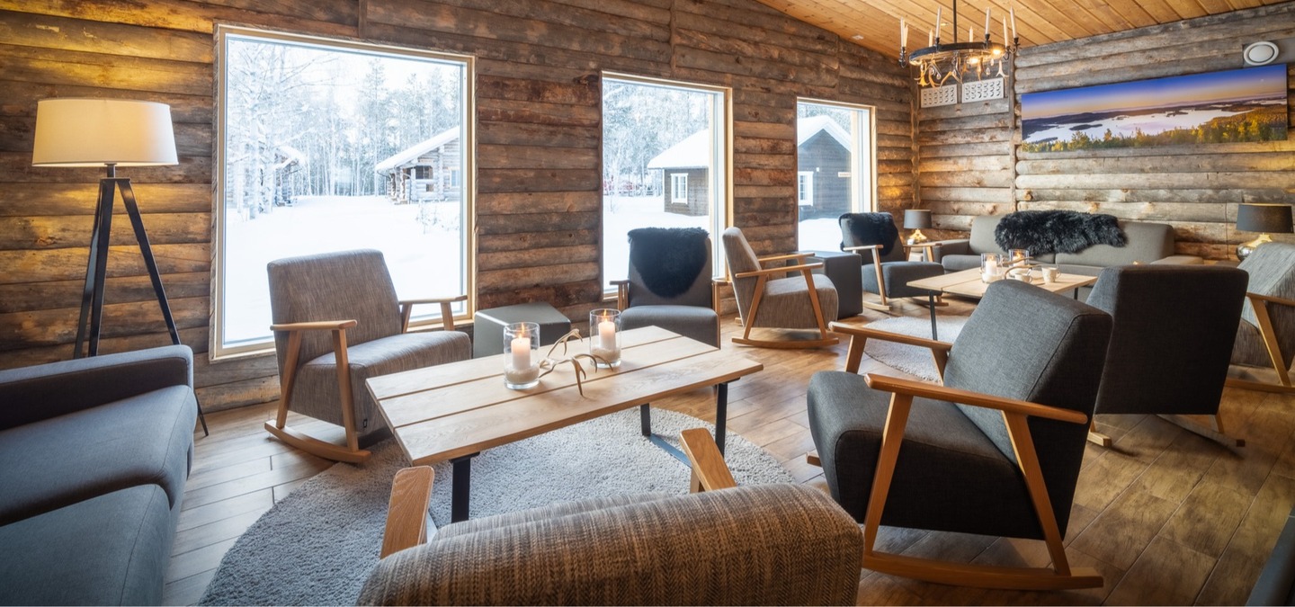 Cozy rustic lounge with log walls, ceiling and large windows overlooking snowy cabins; armchairs, wooden tables and candlelight