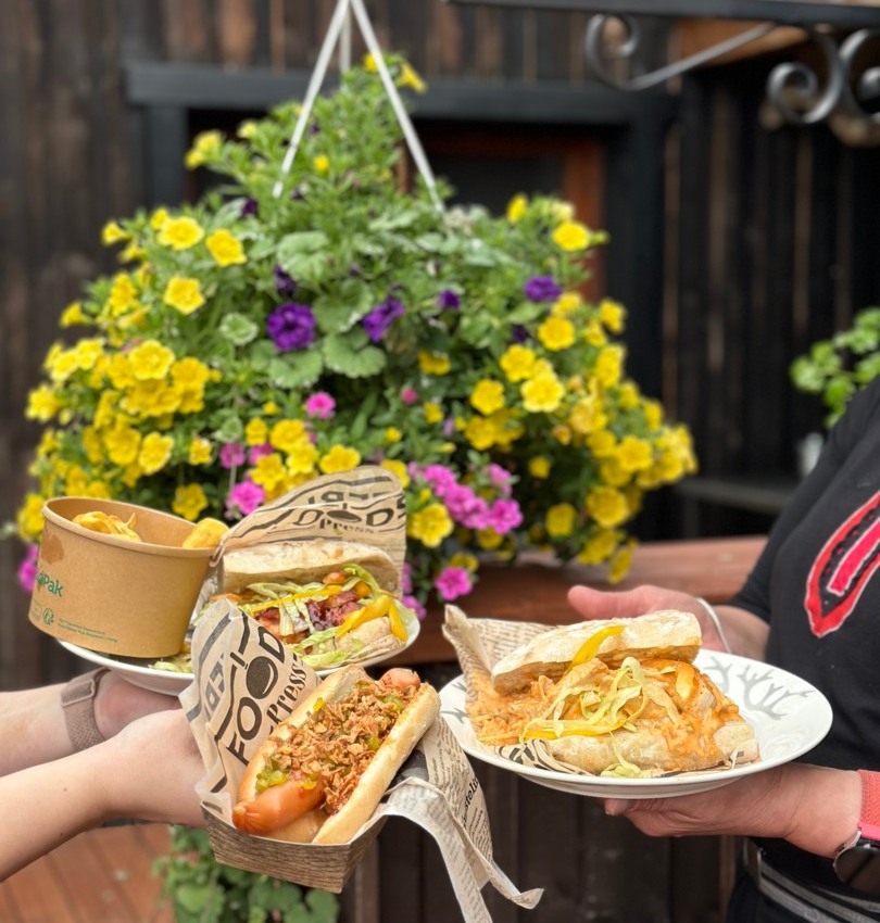 Repas en terrasse décontracté : mains tenant sandwichs et hot-dog croustillant, avec chips, devant une suspension de fleurs jaunes et violettes.