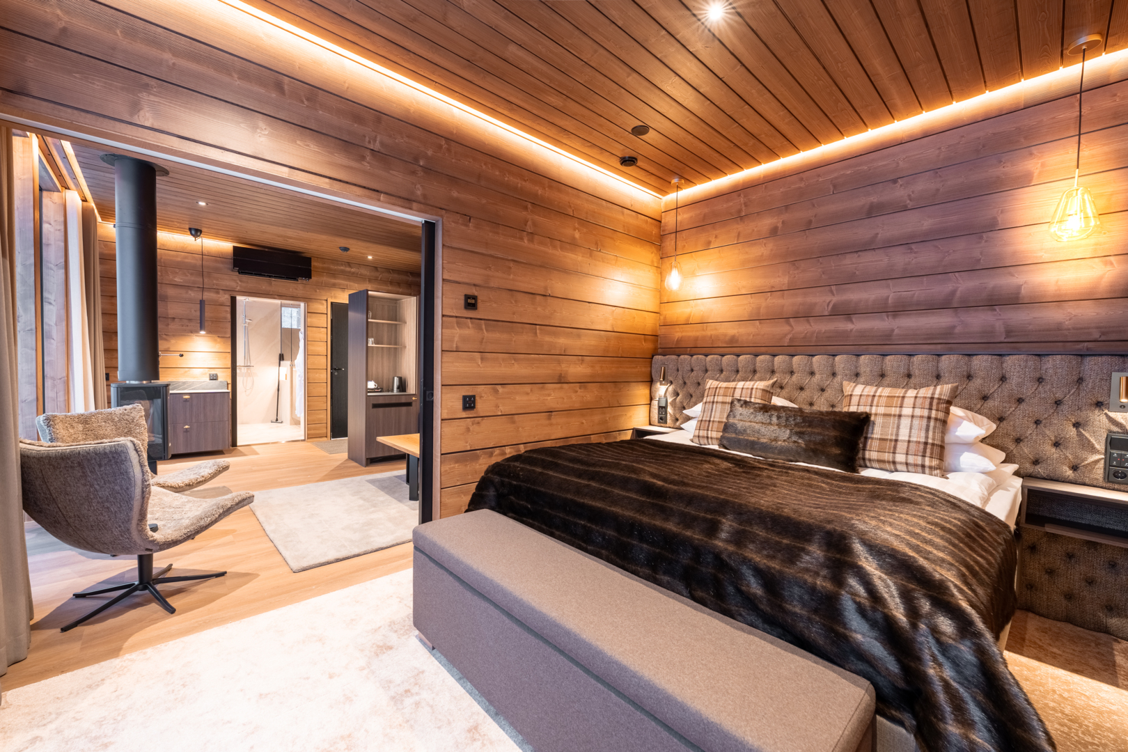 Cozy wood-paneled suite with king bed, tufted headboard, faux-fur throw, plaid pillows, pendant lights and open bathroom.
