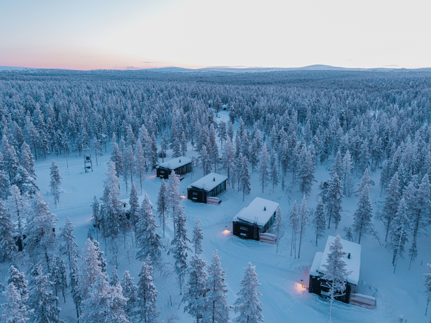 Remote glass-fronted winter cabins with warm entrance lights amid snow-covered pine forest at twilight.