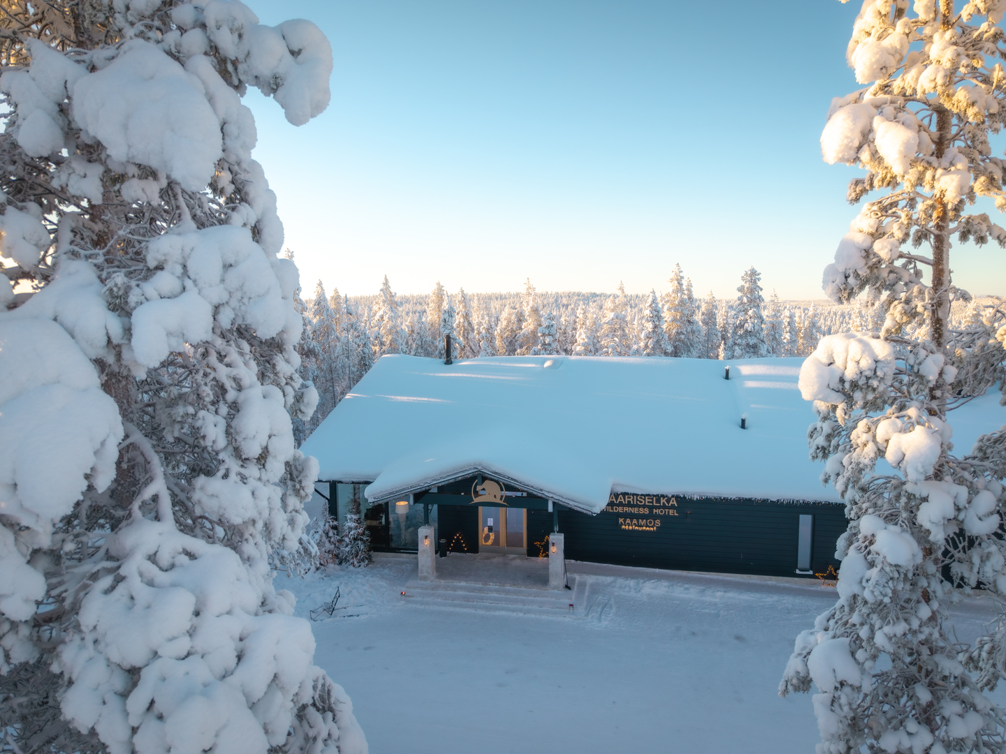 Snow-covered wilderness hotel entrance with heavy rooftop snow, lit doorway and pine trees dusted in soft morning light