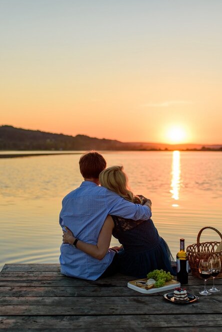 Couple cuddling on a lakeside dock at sunset with picnic basket, wine, cheese, grapes and dessert, tranquil waterfront view