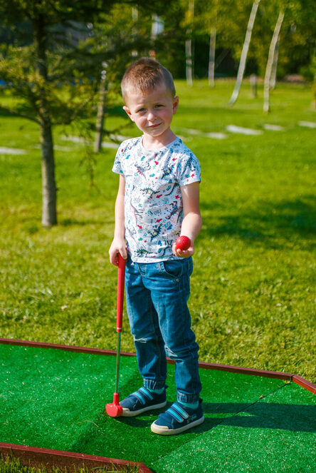 Boy holding red putter and ball on outdoor mini-golf green on a sunny, family-friendly lawn