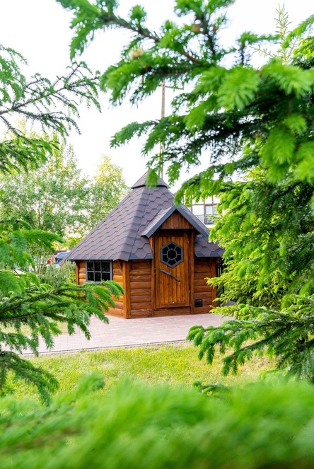 Urokliwy drewniany domek w ogrodzie z dachówką, ośmiokątnymi drzwiami z okienkiem, brukowanym patio, otoczony iglakami