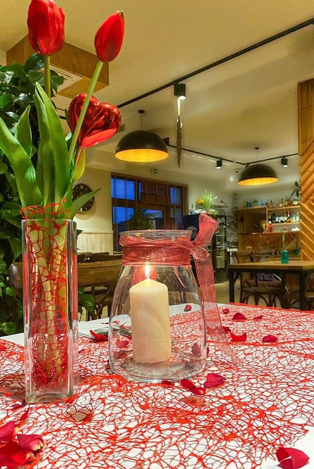 Gemütlicher Restauranztisch mit roten Tulpen im Glas, brennender Kerze im mit Schleife verzierten Glas, Rosenblättern und warmem Licht