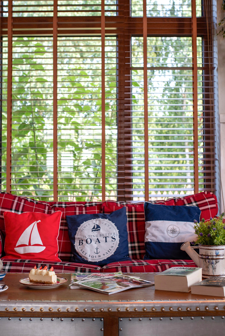 Cozy lounge with red plaid sofas, nautical throw pillows, latte and cake on a trunk-style coffee table, wooden blinds and warm lamp