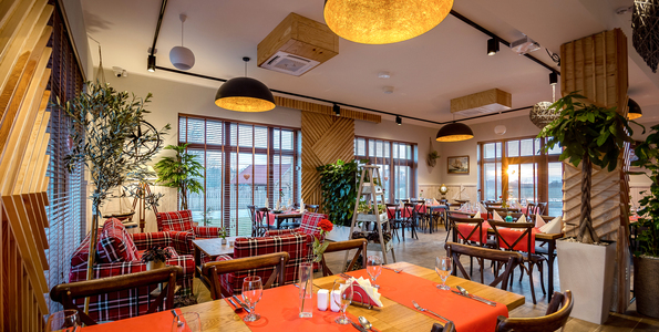 Przytulna restauracja hotelowa z drewnianymi akcentami, wiszącymi lampami, krzesłami w czerwoną kratę, nakrytymi stołami i dużymi oknami.