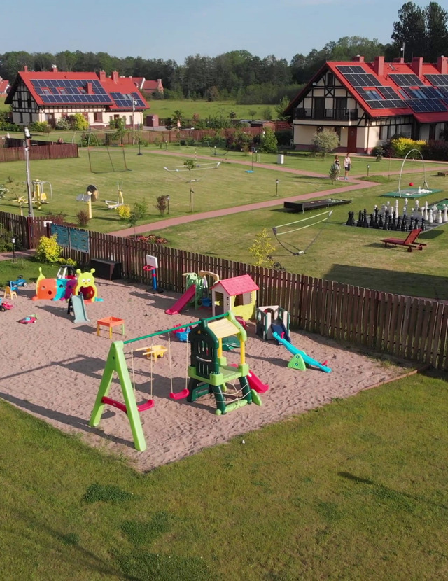 Umzäunter Sandspielplatz mit bunten Rutschen und Schaukeln, daneben Rasen mit Riesenschach, Minigolf, Liegestühlen und roten Cottages mit Solarpaneelen