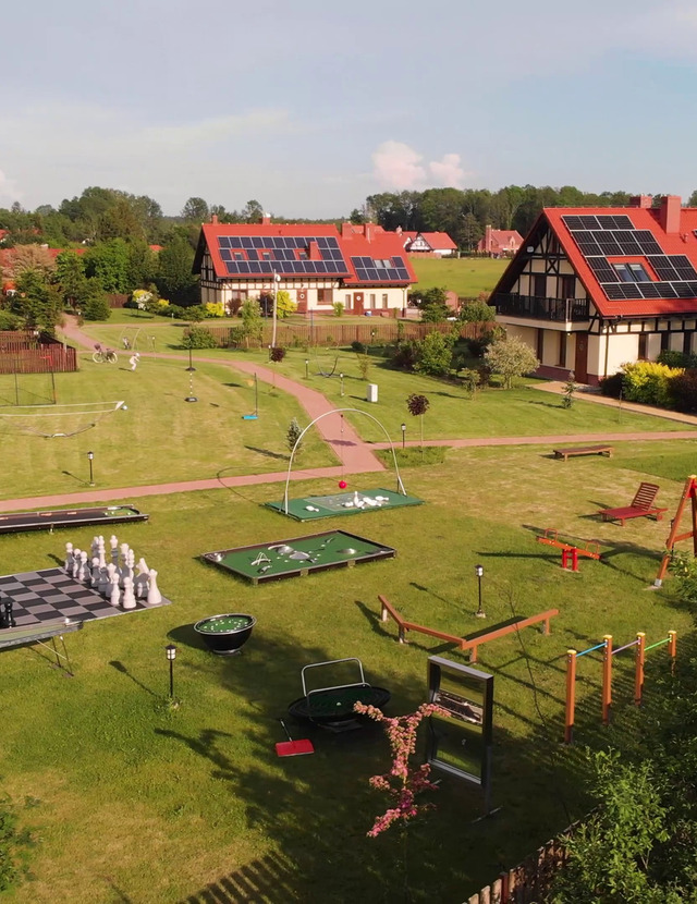 Gästehäuser mit roten Dächern und Solarpaneelen, weitläufige Wiese mit Riesenschach, Minigolf, Spielplatz, Liegen und Spazierwegen.