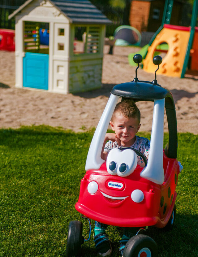 Kind im roten Spielauto auf grünem Hotelrasen, mit Spielhaus und bunter Rutsche im Sandbereich.