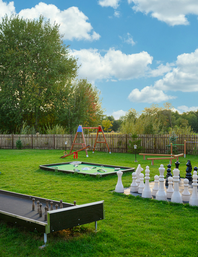 Außen-Spielwiese des Hotels mit übergroßem Schach, Minigolfbahnen, Schaukel und Rasenboccia vor Bäumen und Holzzaun