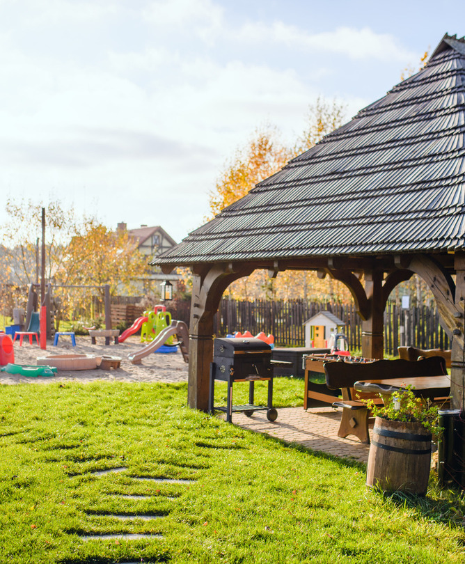 Holzpavillon mit Schindeldach, Außenbank und Grill auf grünem Rasen, daneben bunter Spielplatz für Kinder