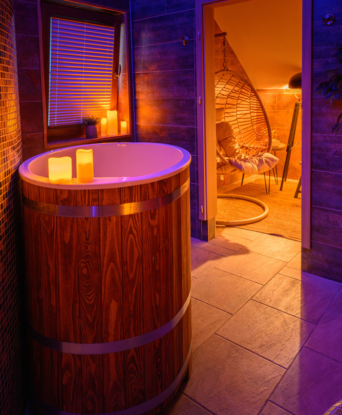Holzbadetonne mit Kerzen im gefliesten Spa, warm beleuchteter Ruhebereich mit hängendem Rattansessel