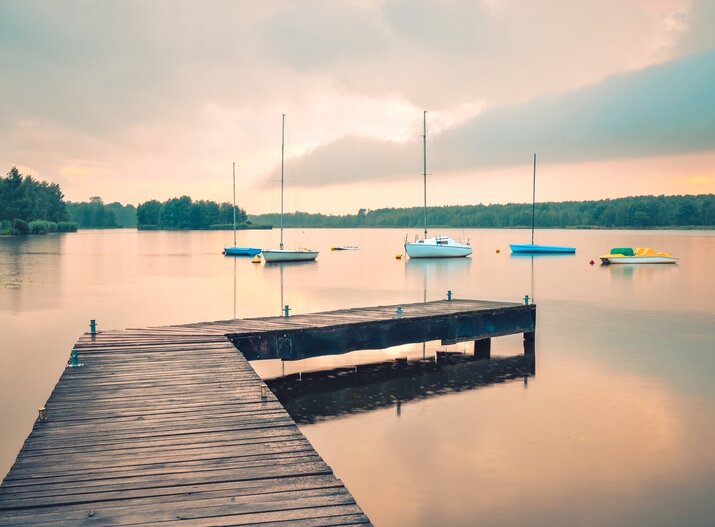 Holzsteg am See bei Sonnenuntergang, vertäute Segelboote, spiegelglattes Wasser und bewaldetes Ufer — ideal für ruhige Wasserurlaube