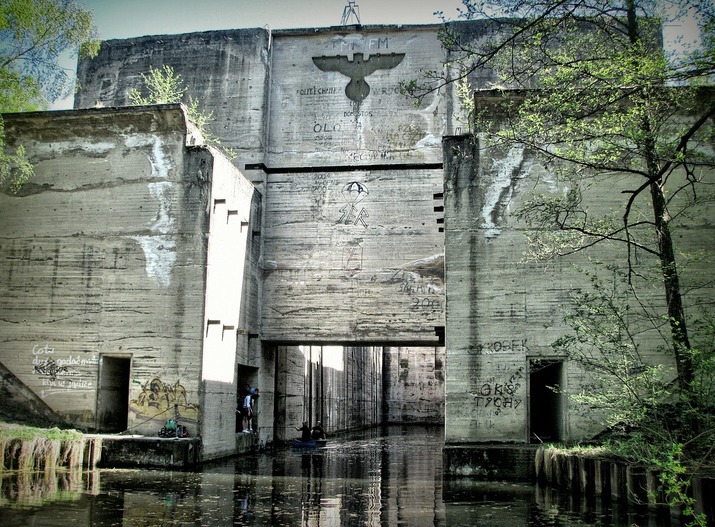 Massive concrete bunker-like structure above a calm canal, graffiti on the walls, trees and people beside a small boat.