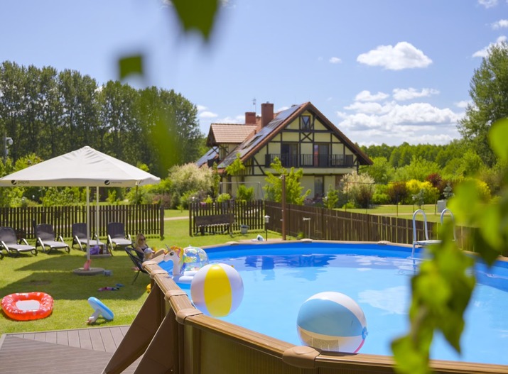Aufstellpool mit Strandbällen und aufblasbaren Spielsachen, Liegen und Sonnenschirm auf dem Rasen, chaletartiges Gästehaus und Garten
