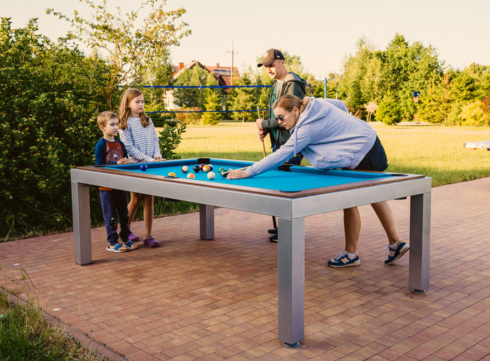 Billardtisch im Freien auf der Hotelterrasse; ein Erwachsener spielt, zwei Kinder und ein weiterer Erwachsener schauen auf dem Rasen zu.