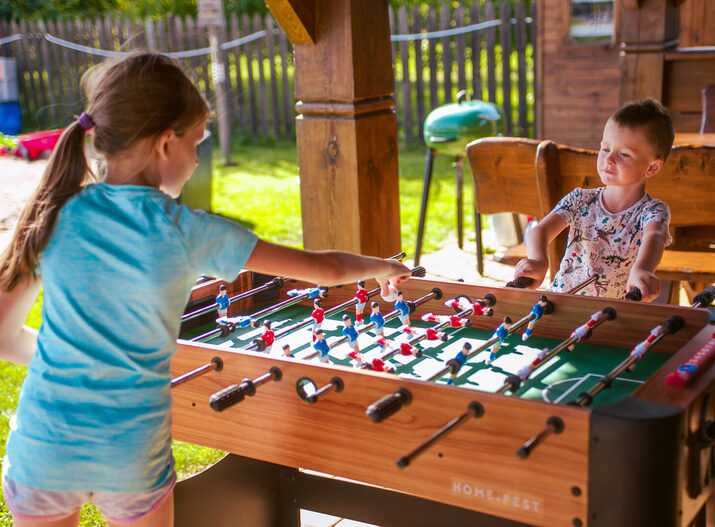 Children playing foosball at a shaded outdoor wooden terrace with benches, lawn and a BBQ grill — family-friendly recreation.