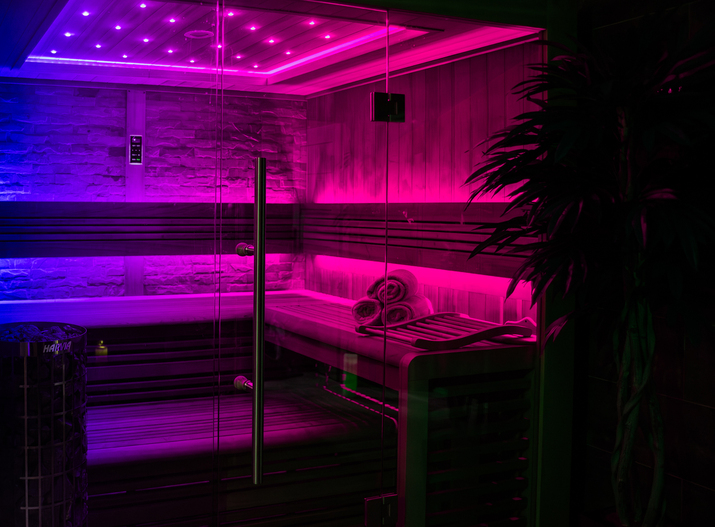 Moderne Sauna mit Glasfront, Holzliegen, stimmungsvollem LED-Licht in Pink und Blau, Ofen mit Saunasteinen und aufgerollten Handtüchern
