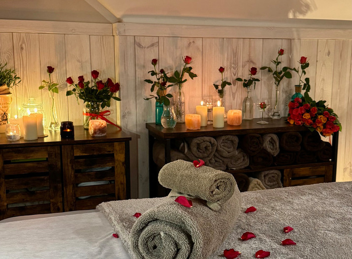 Przytulne studio spa z ręcznikami zwiniętymi na stole do masażu, płatkami róż, zapalonymi świecami i wazonami z czerwonymi różami.