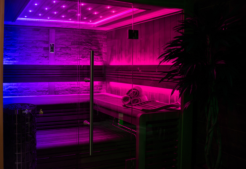 Moderni sauna su stikline priekyne, mediniais suolais, rausvai–mėlynu LED apšvietimu, krosnimi su akmenimis ir susuktomis rankšluostėmis