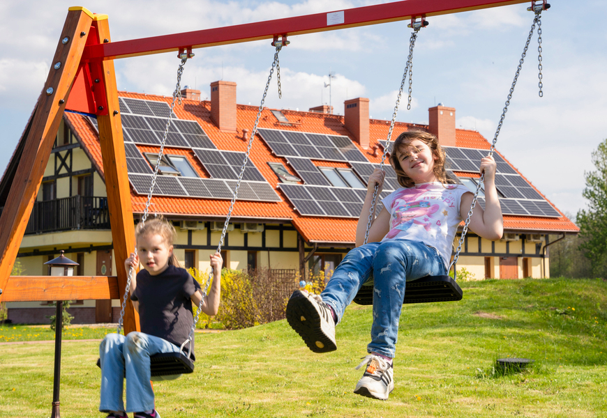 Children on swings in front of a chalet-style hotel with red tiled roof and solar panels, sunny family-friendly lawn