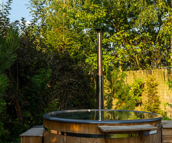 Outdoor wood-fired hot tub on a patio with wooden steps and bench, secluded garden setting framed by tall trees and a bamboo screen