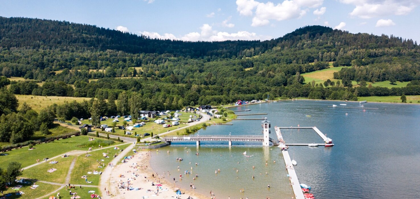 Plaża nad zalewem z pomostem, otoczona zielonymi wzgórzami, z miejscem na piknik i wodnymi atrakcjami
