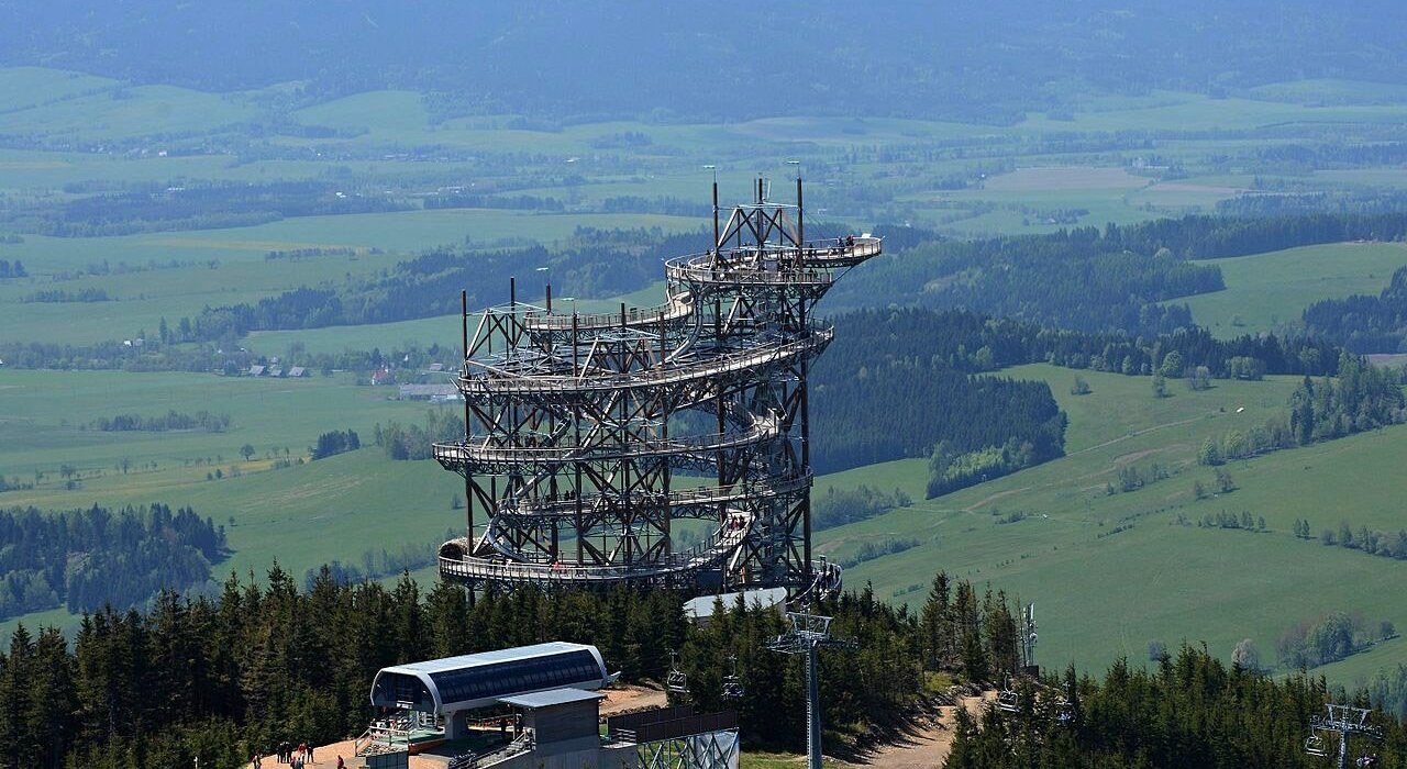 Wielopoziomowa wieża widokowa z tarasem na szczycie w otoczeniu lasu i rozległych zielonych pól w górach