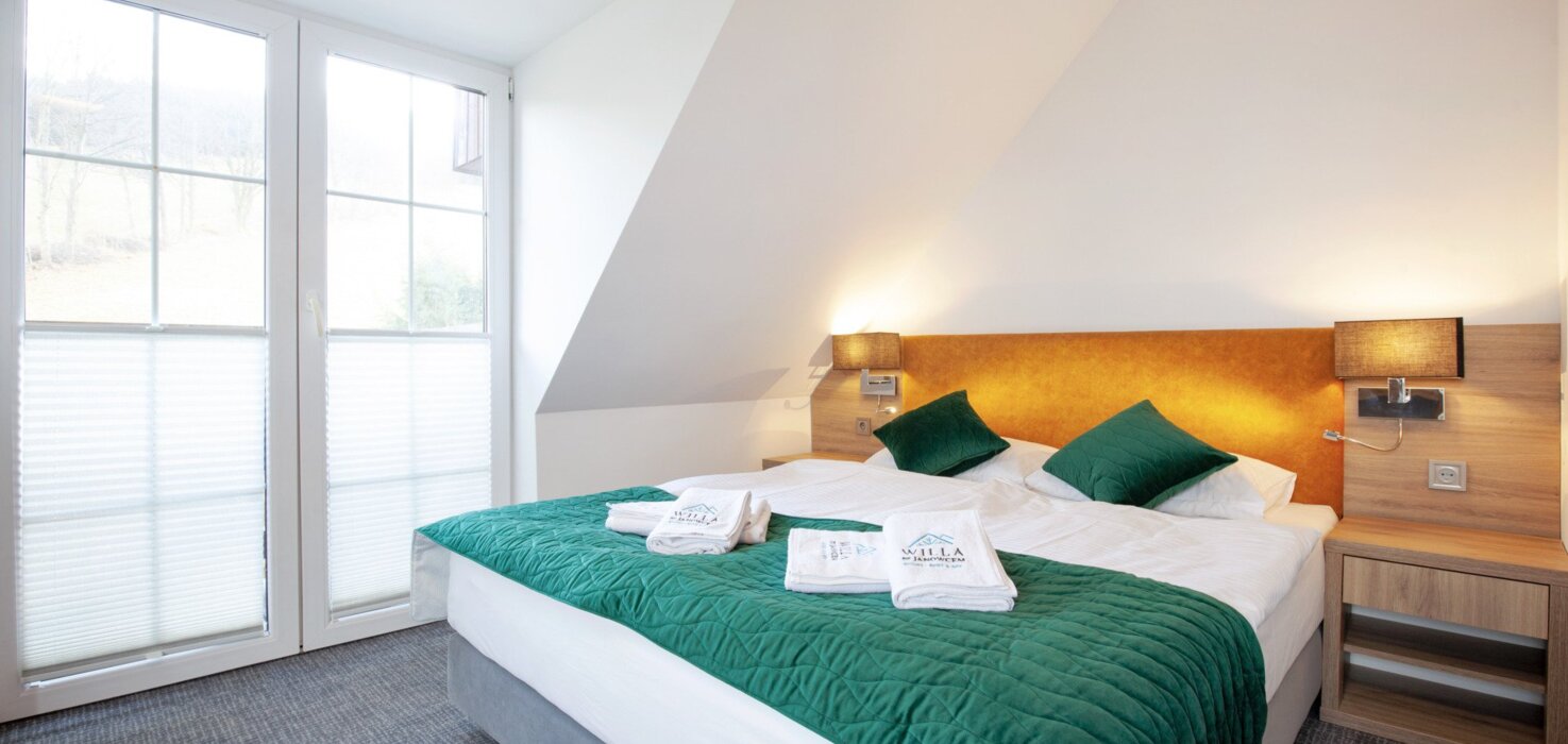 Jasny pokój hotelowy z dużym łóżkiem, zielonymi poduszkami i narzutą, drewnianymi nocnymi stolikami i dużym oknem.