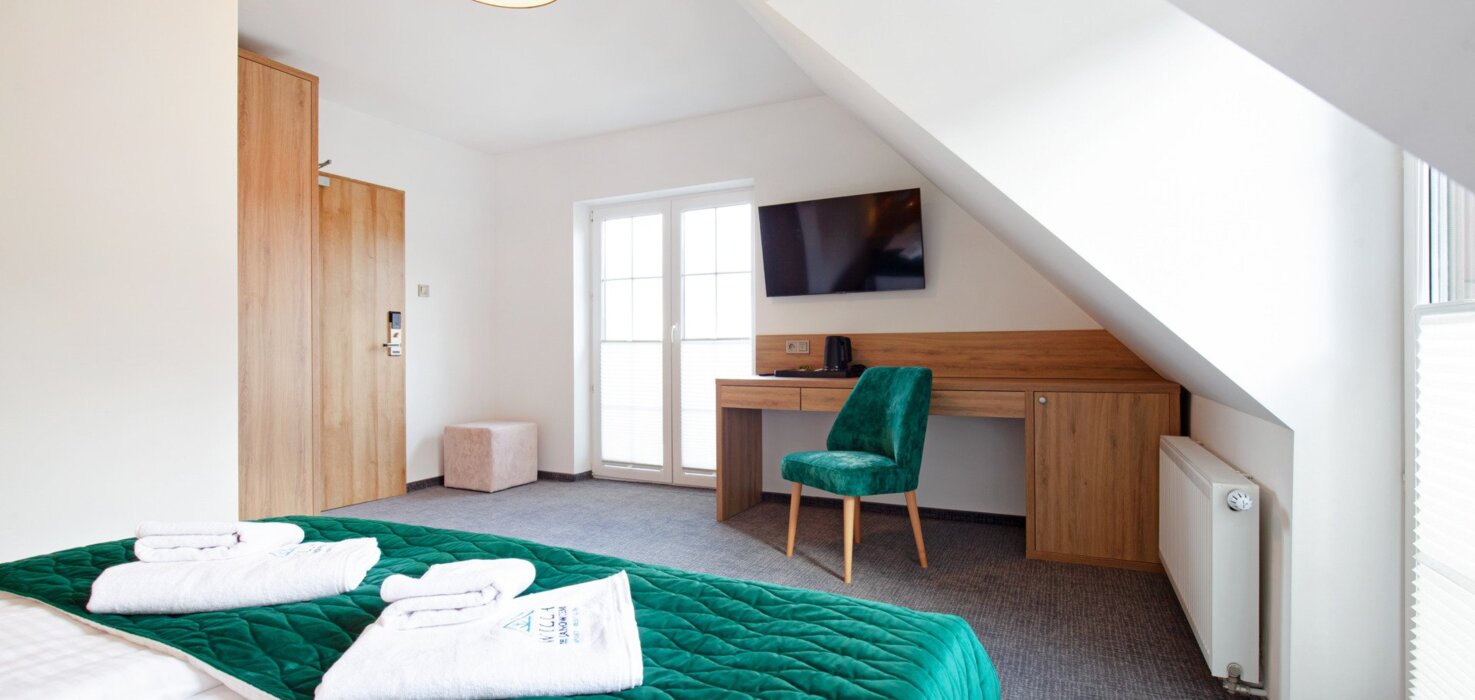 Jasny pokój hotelowy z zielonym narzutem na łóżku, biurkiem, telewizorem na ścianie i miękkim fotelem.