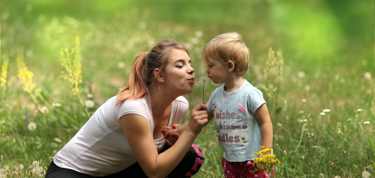 Kobieta z dzieckiem bawią się na łące, dmuchając w dmuchawca w otoczeniu dzikich kwiatów i zieleni.