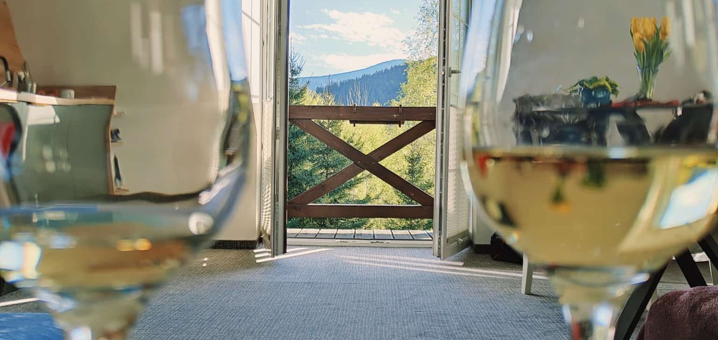 Przytulny pokój z otwartymi drzwiami na balkon z widokiem na góry i las, z dwoma kieliszkami białego wina na pierwszym planie