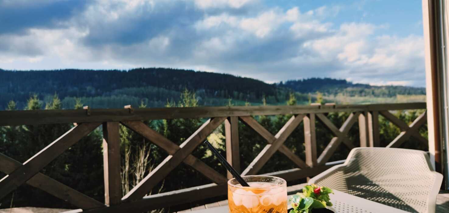 Taras hotelowy z widokiem na zielone wzgórza, stolik z kolorowym napojem i doniczką z kwiatem na tle drewnianej balustrady.