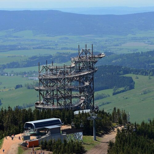 Wielopoziomowa wieża widokowa z tarasem na szczycie w otoczeniu lasu i rozległych zielonych pól w górach