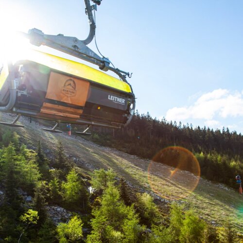 Nowoczesna gondola wyciągu linowego nad zielonym stokiem narciarskim otoczonym lasem sosnowym pod słonecznym niebem