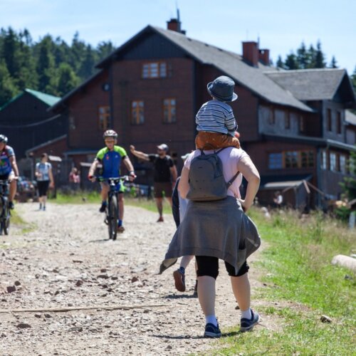 Drewniany górski pensjonat z turystami spacerującymi i rowerzystami na szutrowej ścieżce w słoneczny dzień