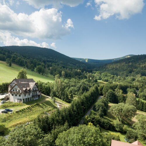 Urokliwy pensjonat z balkonami na wzgórzu otoczony zielonymi lasami i malowniczymi górami w tle pod błękitnym niebem