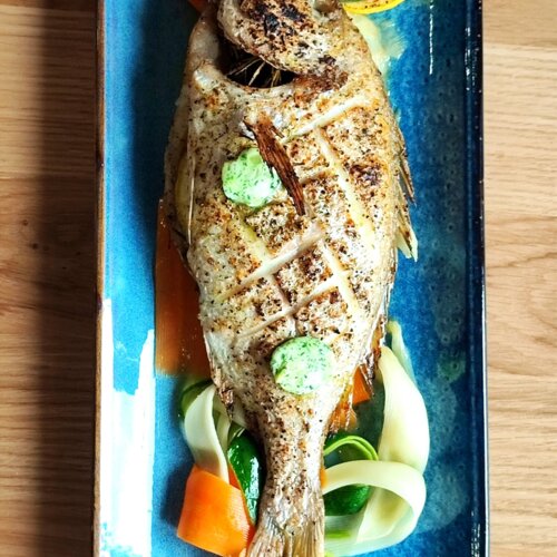 Grillowana ryba podana na niebieskim talerzu z plastrami cytryny i warzywami, doprawiona ziołowym masłem